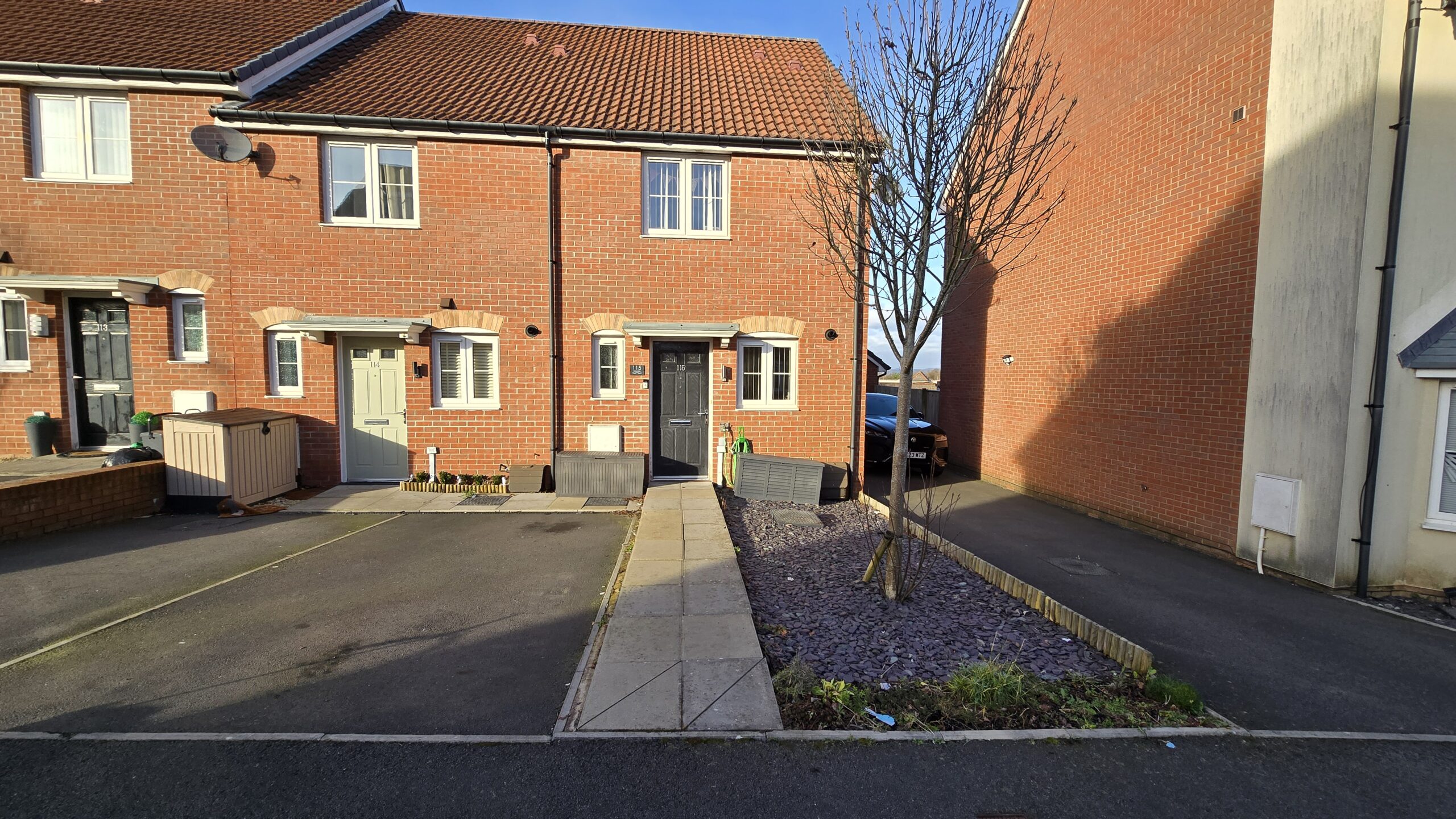 Image of a red brick house with a black front door with a path going to it. On the right side of the path is pebble dash with a tree. To the left of the path is a parking space.